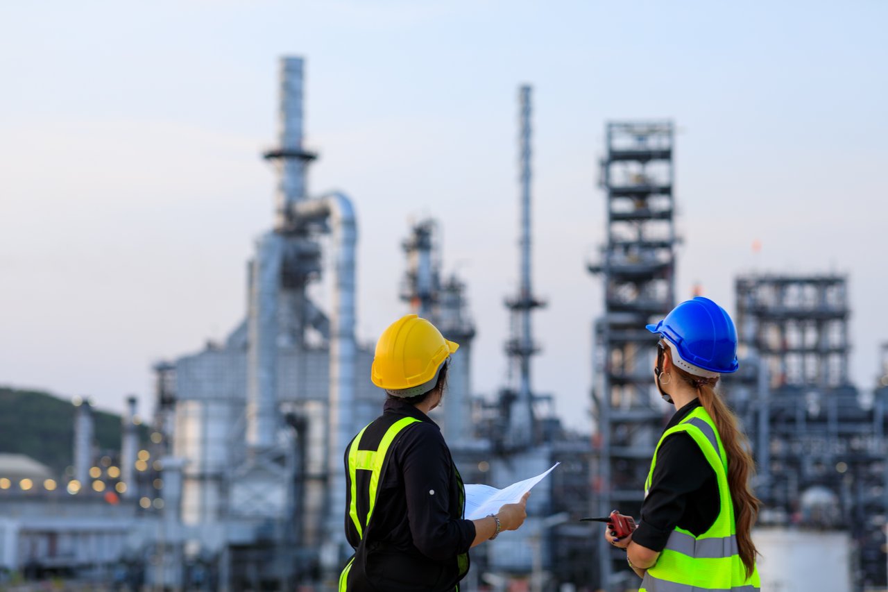 Zwei Arbeiter mit Helmen und Warnweste vor einer Chemiefabrik